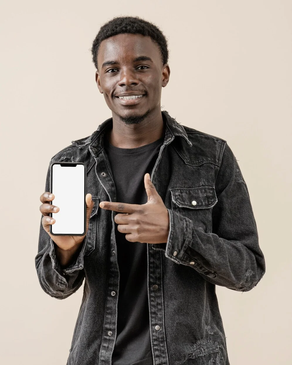 A smiling young man in a denim jacket points at the phone in his hand