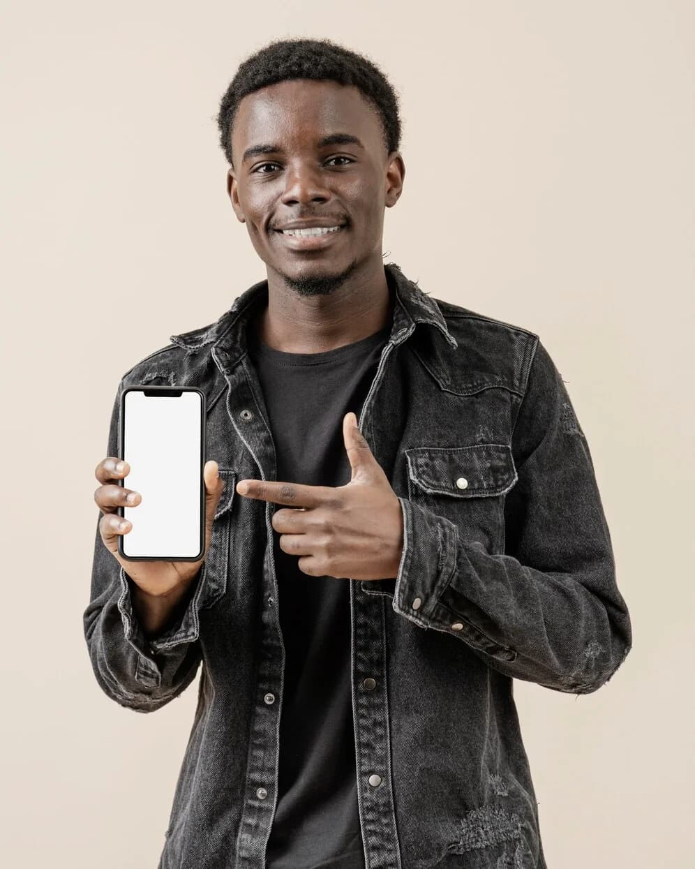 A smiling young man in a denim jacket points at the phone in his hand